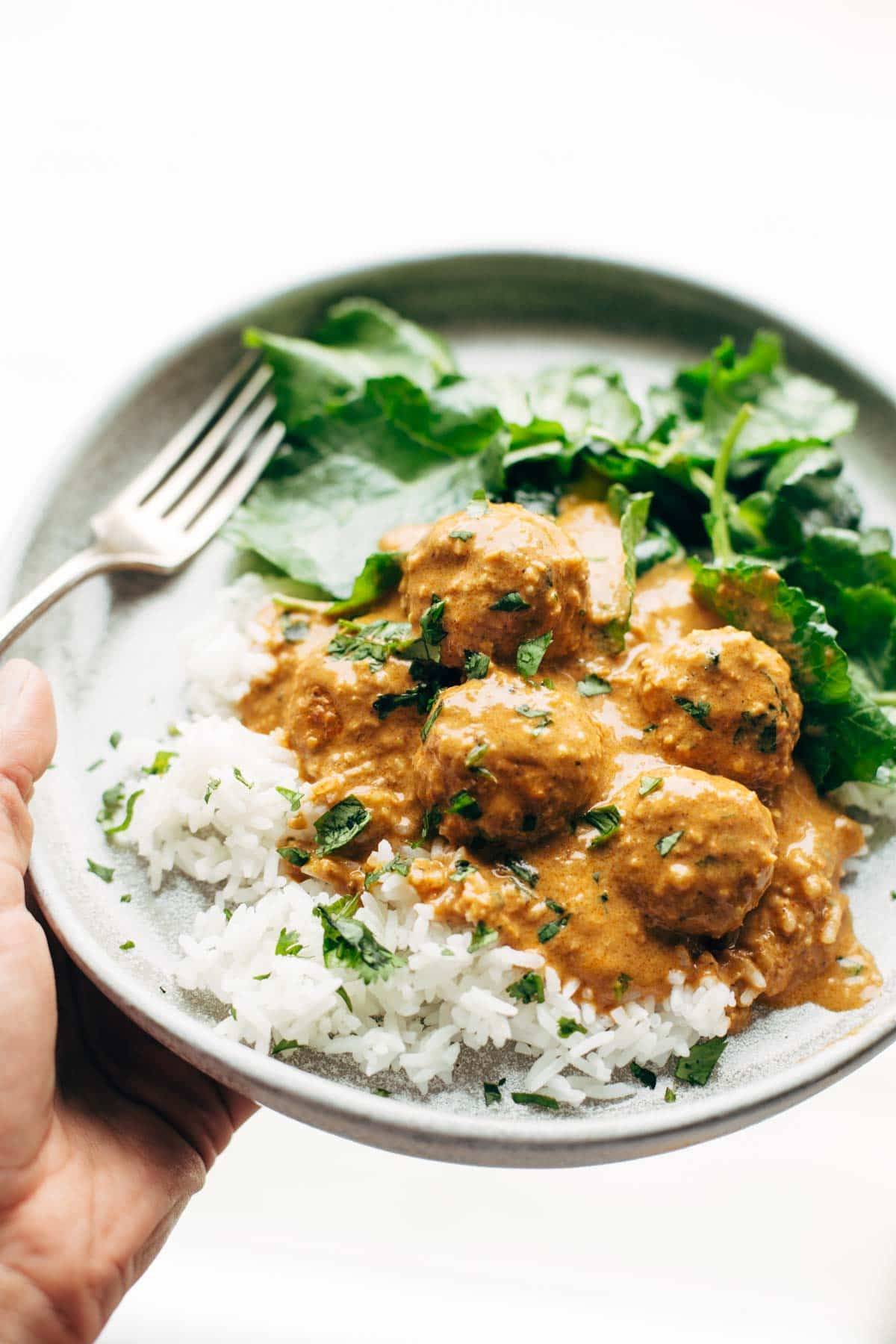 White hand holding a plate with cauliflower vegetarian meatballs, white rice, and fresh herbs sprinkled on top. There is a fork on the plate with greens. 