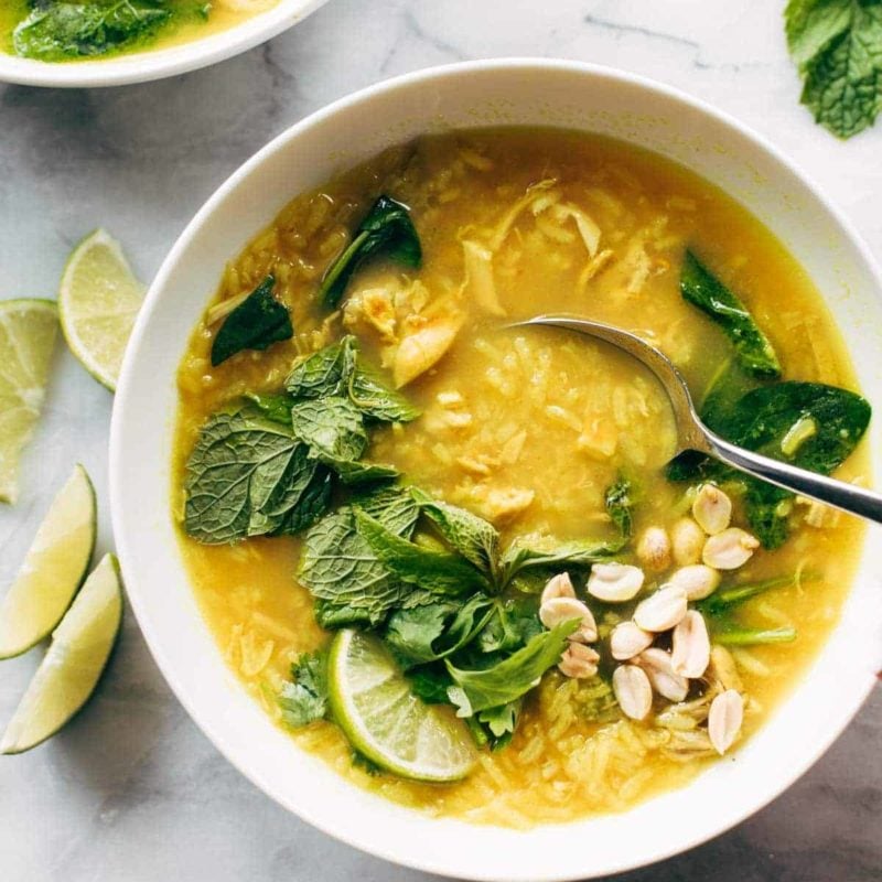 Chicken and rice soup in a bowl with a spoon. There are fresh herbs on top and lime wedges on the side.