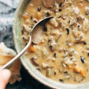 White hand holding a spoon in a bowl of Wild Rice Soup. There's broken crusty bread by the soup bowl.