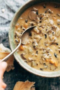 White hand holding a spoon in a bowl of Wild Rice Soup. There's broken crusty bread by the soup bowl.