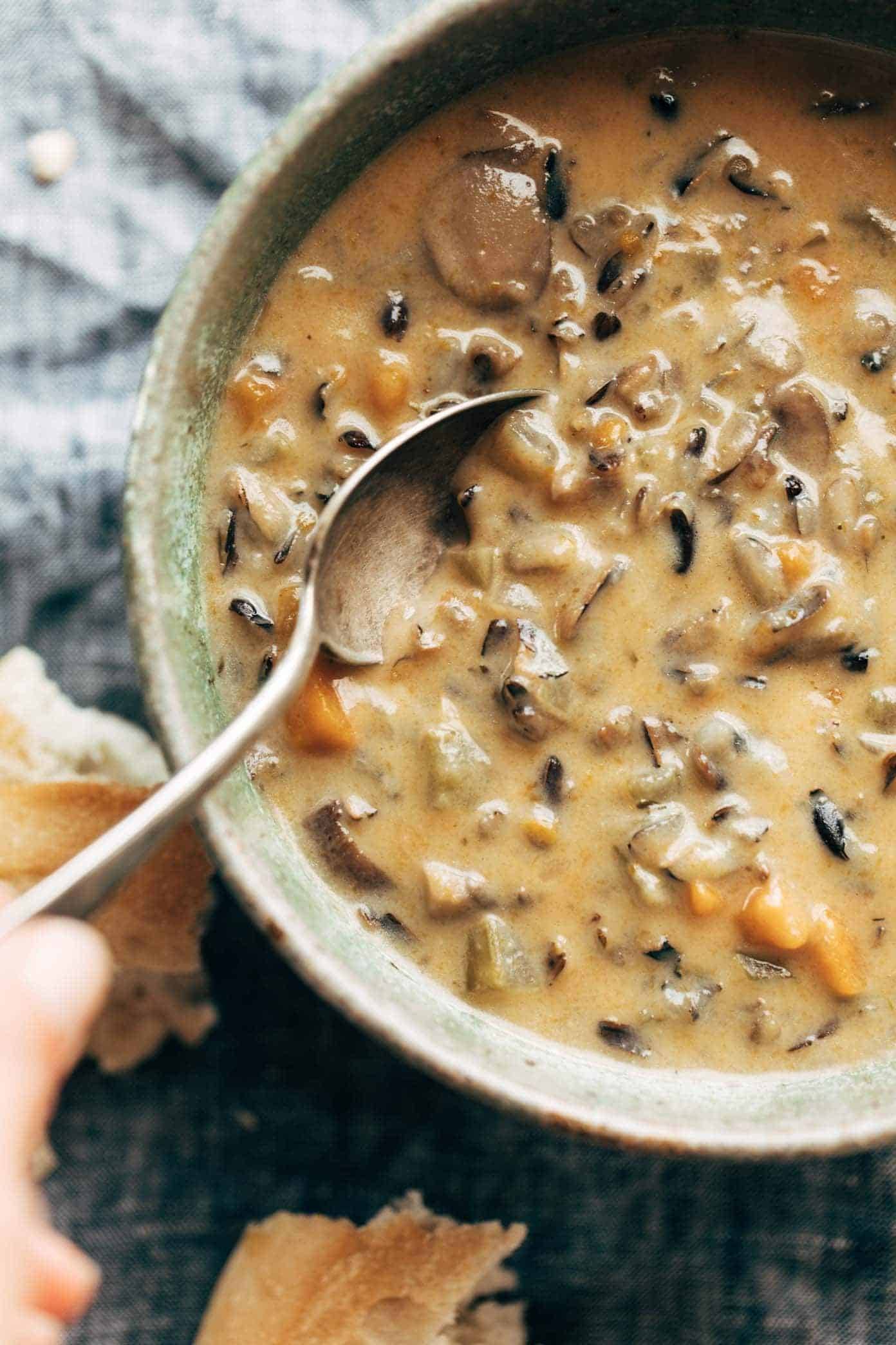 White hand holding a spoon in a bowl of Wild Rice Soup. There's broken crusty bread by the soup bowl. 